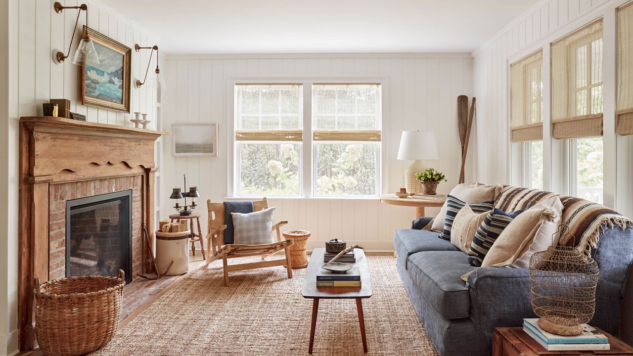 A cozy, coastal-inspired living room featuring white vertical shiplap walls and a rustic brick fireplace with a carved wood mantel. A denim-blue sofa sits on a large jute rug, adorned with neutral and striped pillows. To the left, a light wood sling chair faces a mid-century modern coffee table. Large windows with woven bamboo shades fill the room with natural light, highlighting decorative accents like vintage oars, wicker baskets, and brass sconces framing a seascape painting above the mantel.