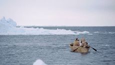 Two indigenous Arctic peoples paddle through icy waters in a traditional canoe-style boat.
