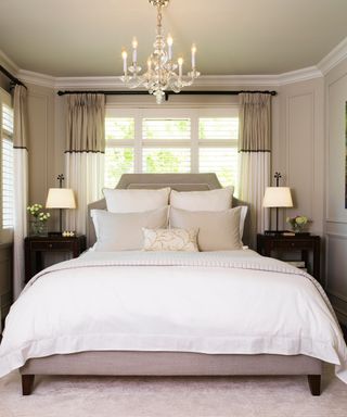 A bedroom with a gray bed with white bedding, a chandelier, and a window with brown and white curtains