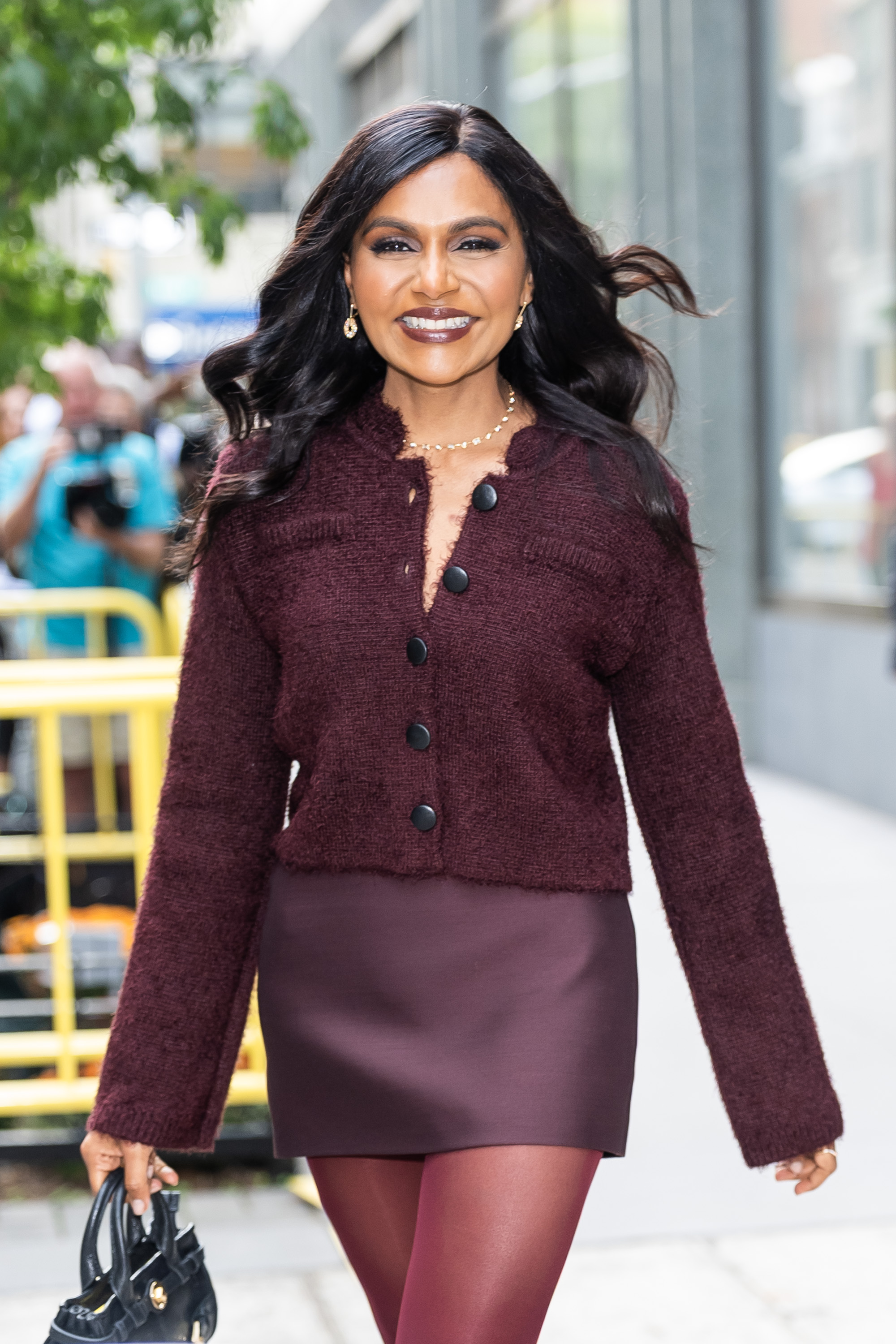 a dark-haired woman photographed by paparazzi wearing a burgundy sweater, a silk, maroon miniskirt, and dark red tights