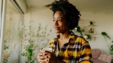 woman holding a cup and looking out fo the window, sideways to the camera. she's wearing a yellow checked shirt with plants behind her in a living room setting. 