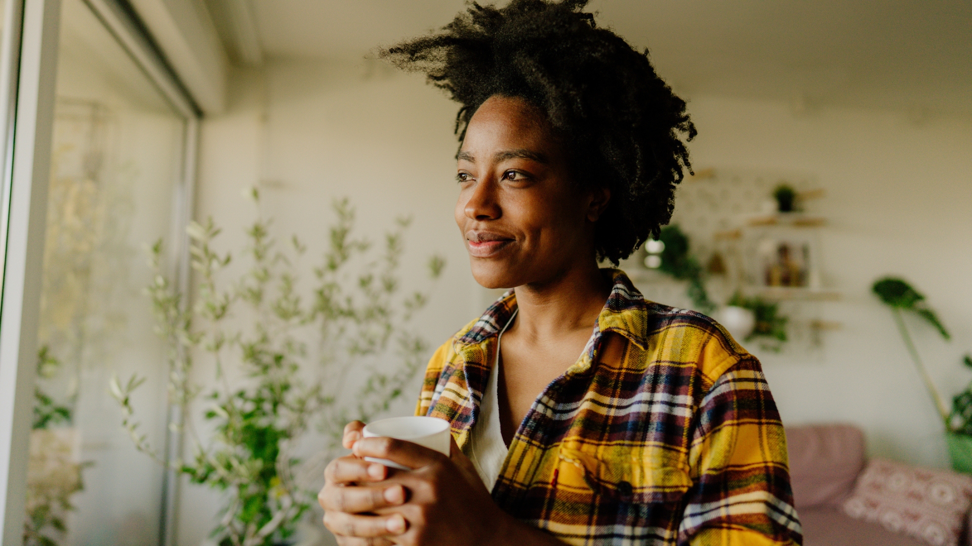 woman holding a cup and looking out fo the window, sideways to the camera. she's wearing a yellow checked shirt with plants behind her in a living room setting.