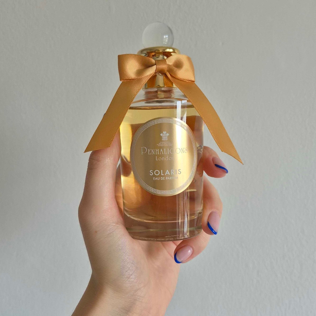 A close-up of Digital Beauty Writer Naomi Jamieson's hand, seen holding a bottle of Penhaligon's Solaris perfume, which features an orange bow/ in front of a cream wall background