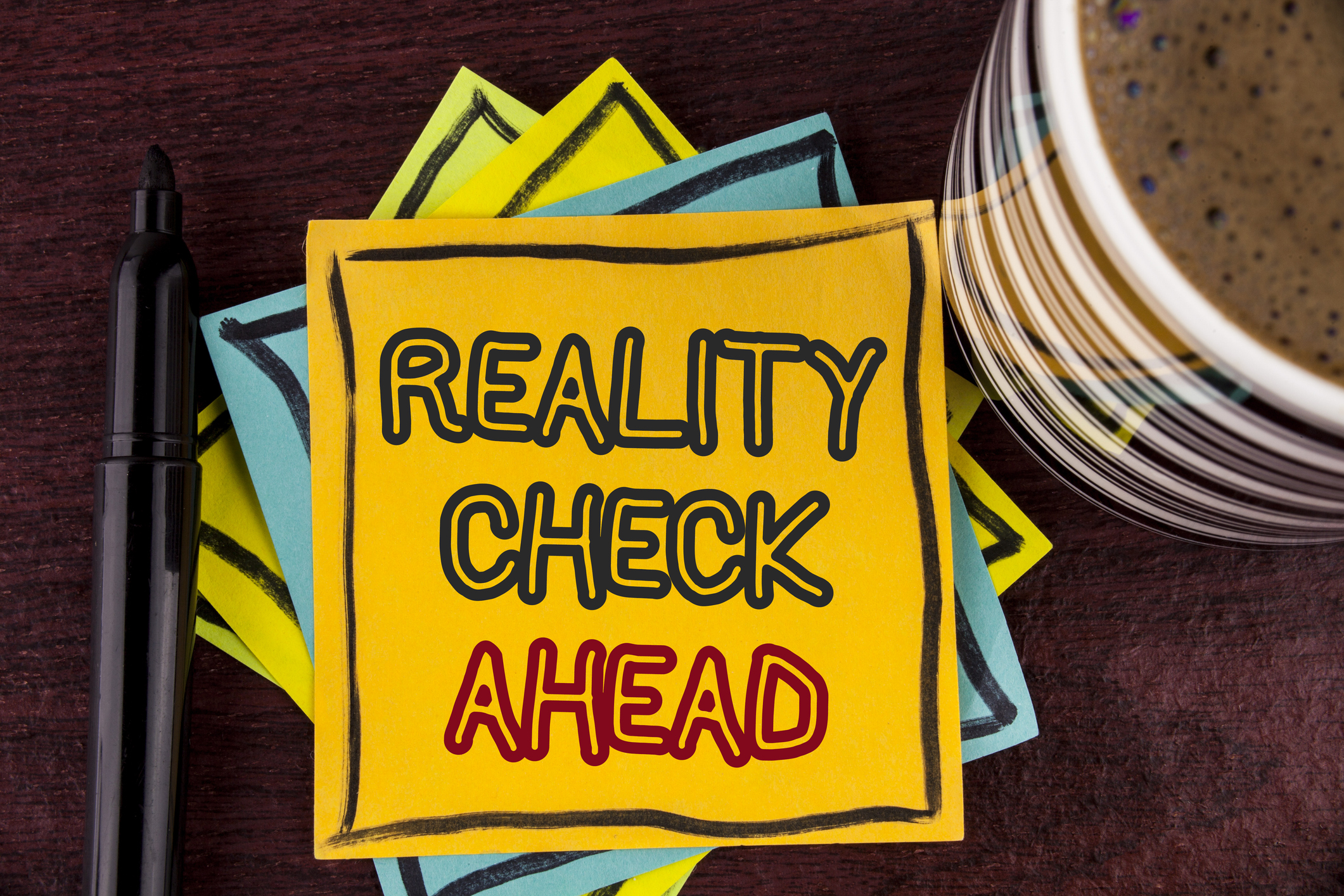 stack of Post-it notes with a yellow one on top that reads "reality check ahead" next to a coffee mug