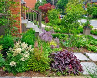 This urban front yard garden features a large veranda, brick paver walkway, retaining wall with plantings of bulbs, shrubs and perennials.