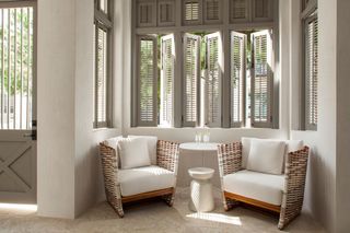Vignette in a room with two rattan armchairs, a small white side table, white walls and warm grey shutters