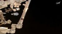 a white space capsule is seen against the darkness of space, with parts of the international space station visible in the foreground