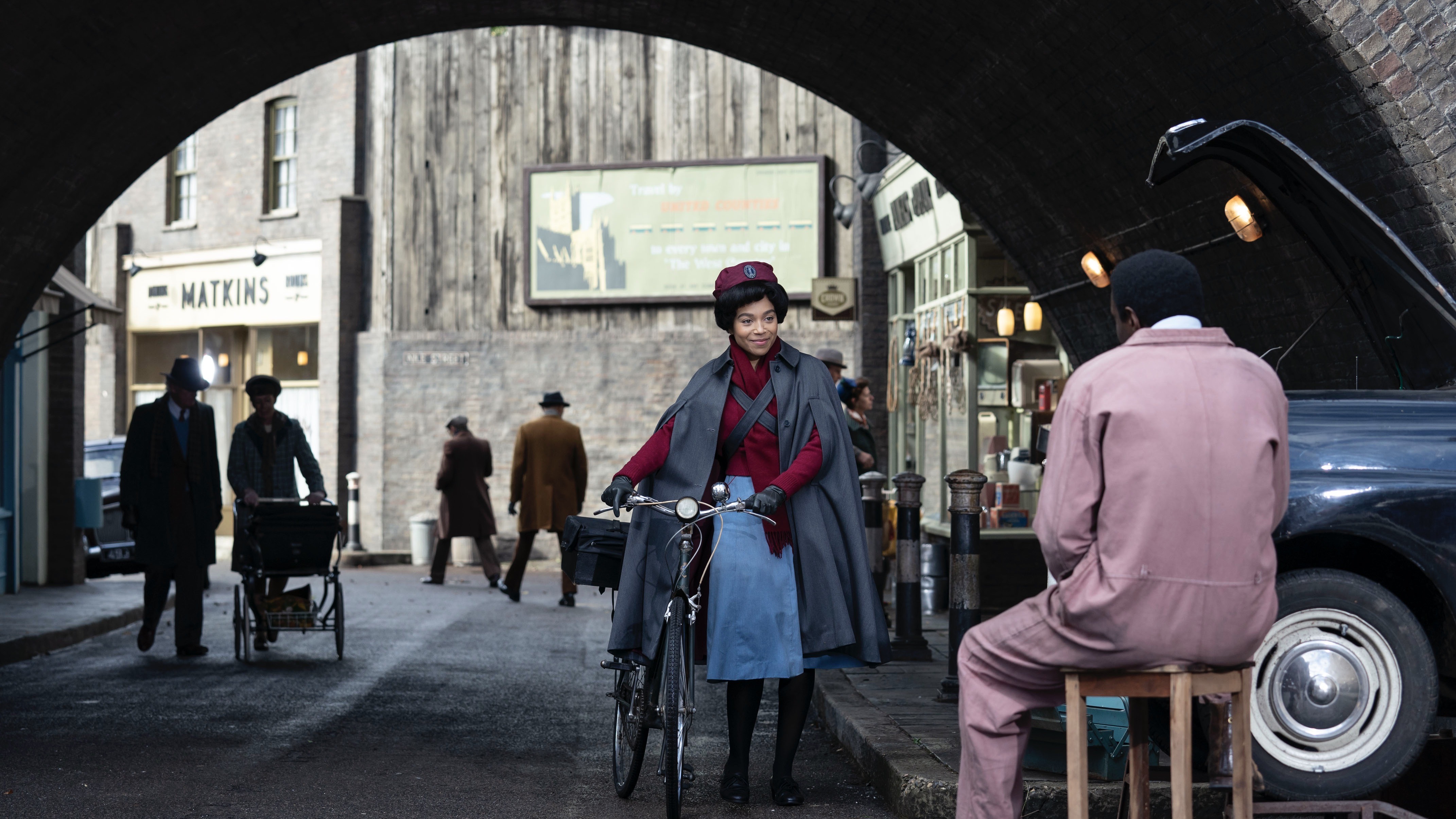 Leonie Elliott in nurse's uniform as Lucille pushes a bike towards Zephryn Taitte in overalls as Cyril in Call the Midwife.