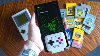 Hand holding Razer Edge with 8Bitdo FlipPad Game Boy controller inserted into the bottom with DMG Game Boy and cartridges on table below.