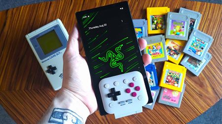 Hand holding Razer Edge with 8Bitdo FlipPad Game Boy controller inserted into the bottom with DMG Game Boy and cartridges on table below.