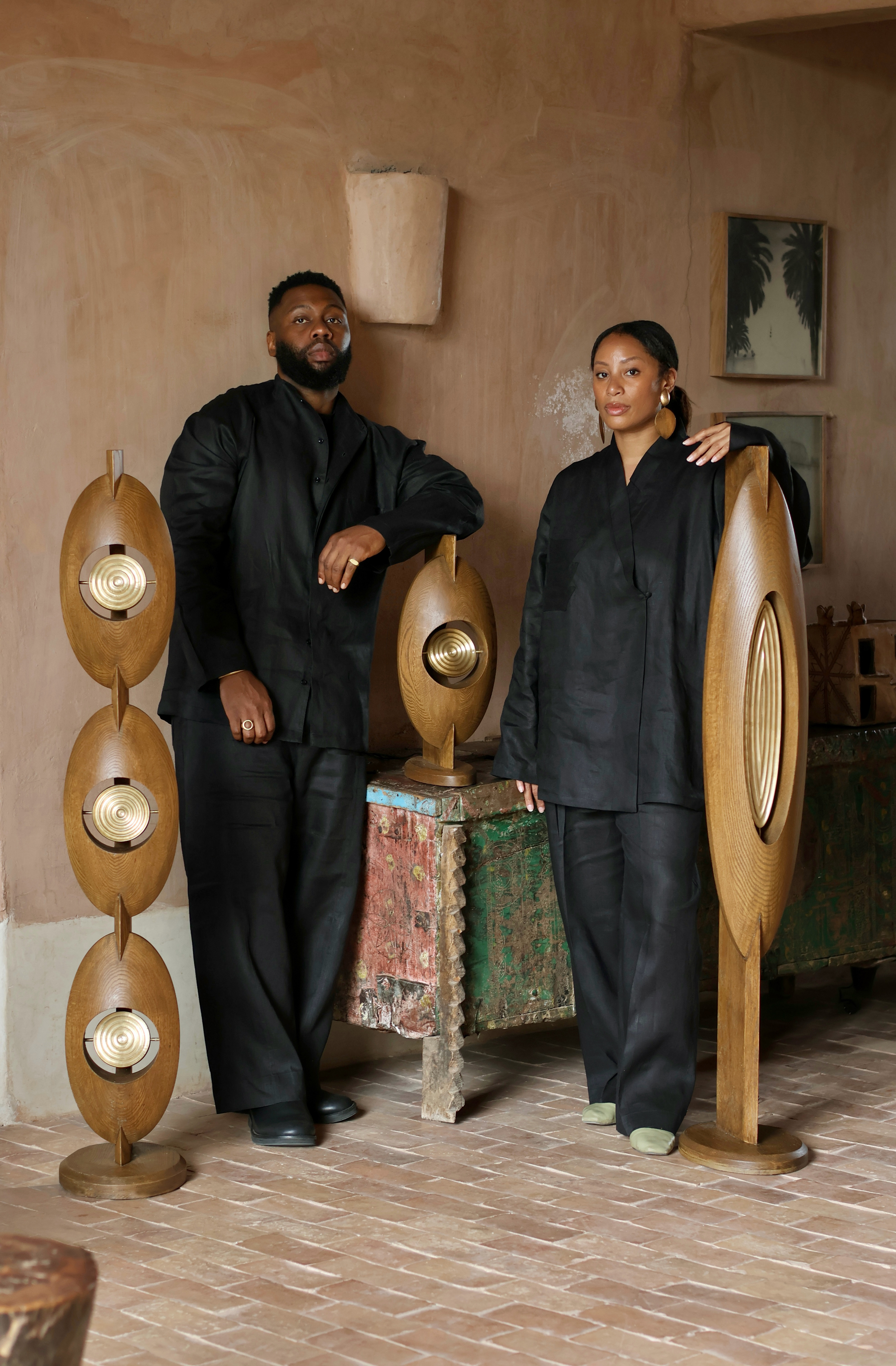 A male and female designer, dressed in total black look, stand next to three totemic wooden sculptures in a terracotta-clad room with distressed wood furniture and abstract artworks.