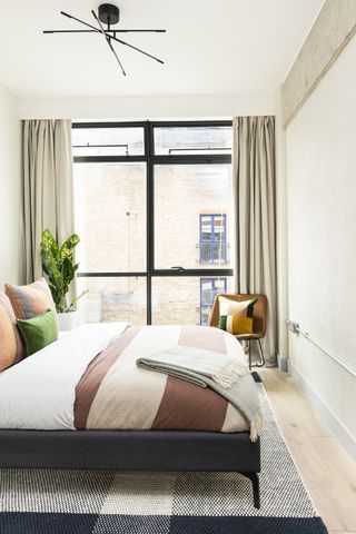 small modern bedroom with black bed, neutral scheme, black pendant light, chair in corner, check rug, cream drapes, stripe blanket, stripe cushions, plant