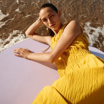 Model wearing a yellow &OtherStories dress lying on a pink board on the sea shore