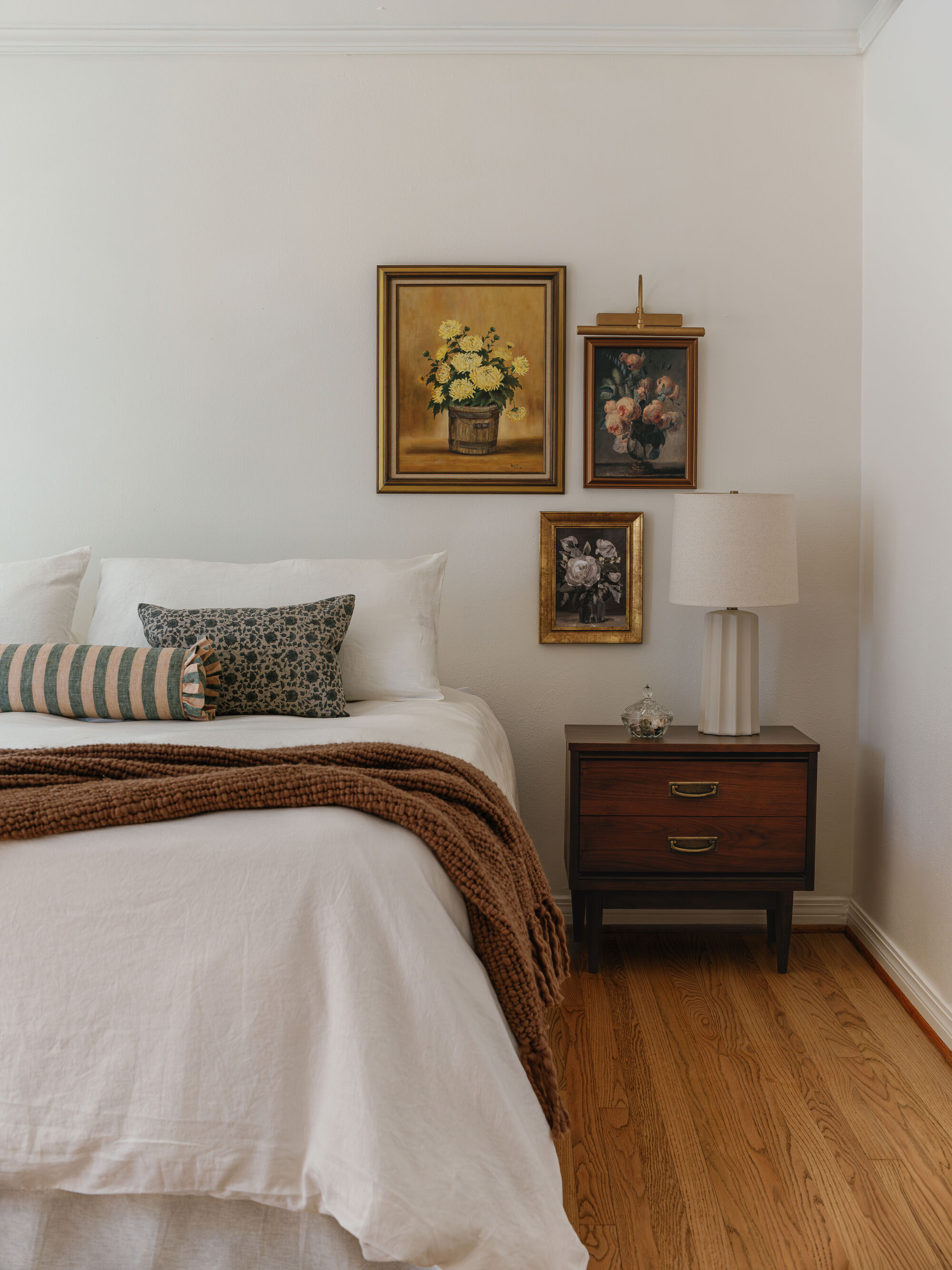 A white bedroom with white bedding and textured throws and pillows. There is a wooden side table with a white lamp and three artworks above the side table.