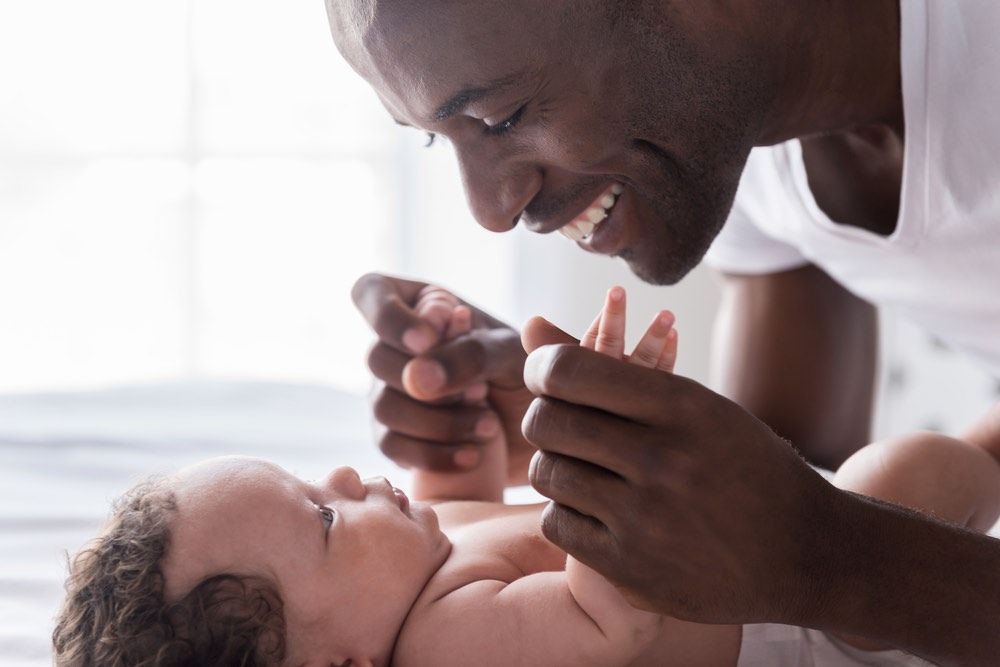 Side view of happy young African man playing with his little baby and smiling while lying in bed.