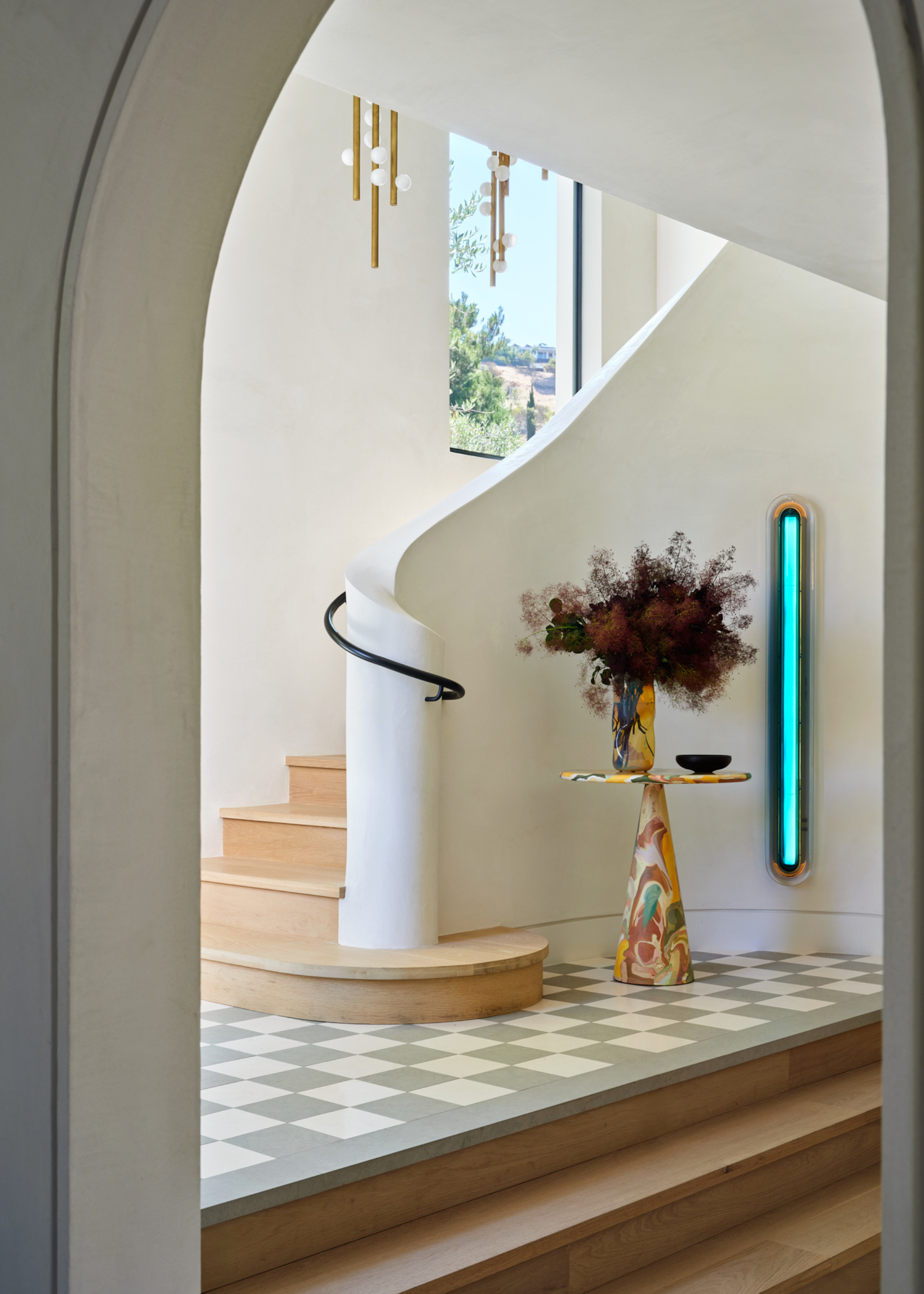 A bright entryway with a curved white staircase, checkerboard tiled floors, a colorful side table with a vase of fluffy stems and a blue wall light