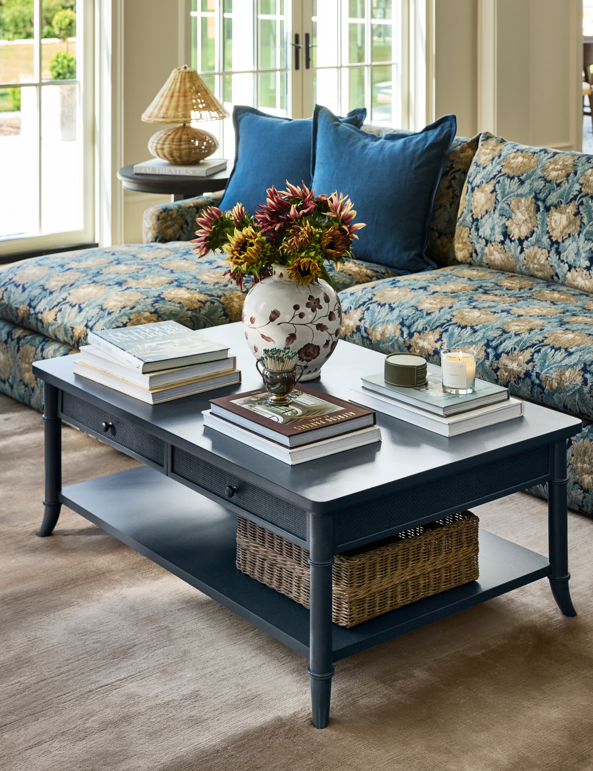 Two-tier navy blue coffee table with storage styled with stacks of coffee table books, a floral-patterned vase, match strike, and lit candles.