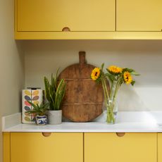 a yellow kitchen with white worktops, a vase of sunflowers and a large round wooden chopping board