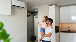 male holding young female child in his arms, with child holding remote control pointing it at an air con unit on wall in kitchen area