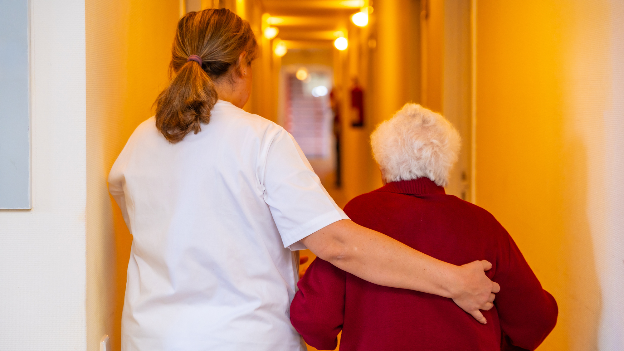 A woman with short blond hair wearing a white shirt escorts an older woman with short white hair and a red sweater down a hallway.