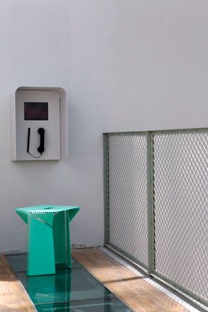 A green armless chair next to a grey public wall phone, photographed during the day