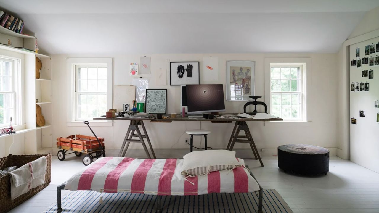 A white home office-lounge with a large wooden desk with a monitor and home office essentials on it, two windows either side of the desk, a small sofa bed in the middle, and built-in storage to the left around the window