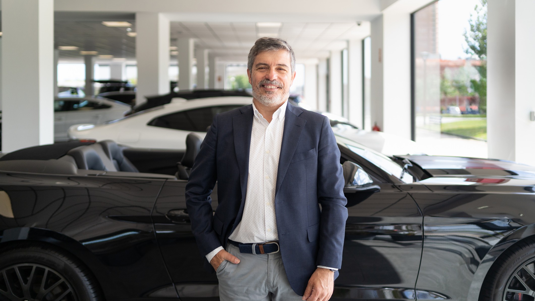 A man in business casual stands in front of a shiny car at a dealership. 