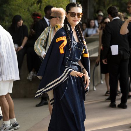 A woman wears a navy letterman jacket and a denim dress with large sunglasses.