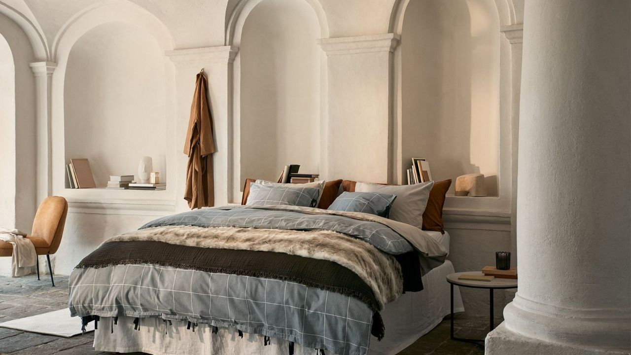 A Renee Kemps bedroom: a large bed with blue and beige bedding against white walls.