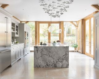 Large white kitchen space with statement marble island, wooden joinery and door frames, large stone floor tiles, white kitchen cabinets, decorative hanging feature and lights over island