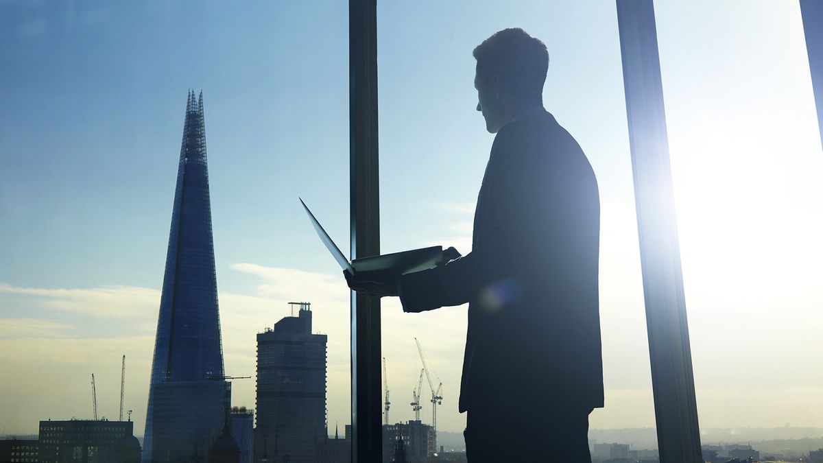 A businessman holding a laptop looking out the window at a London skyline including The Shard skyscraper