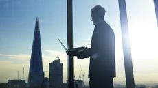 A businessman holding a laptop looking out the window at a London skyline including The Shard skyscraper