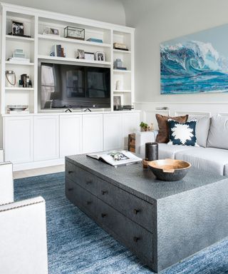 Bright white living room with white storage and display unit with tv, large gray coffee table with draws, blue rug and blue artwork on the wall with a wave design