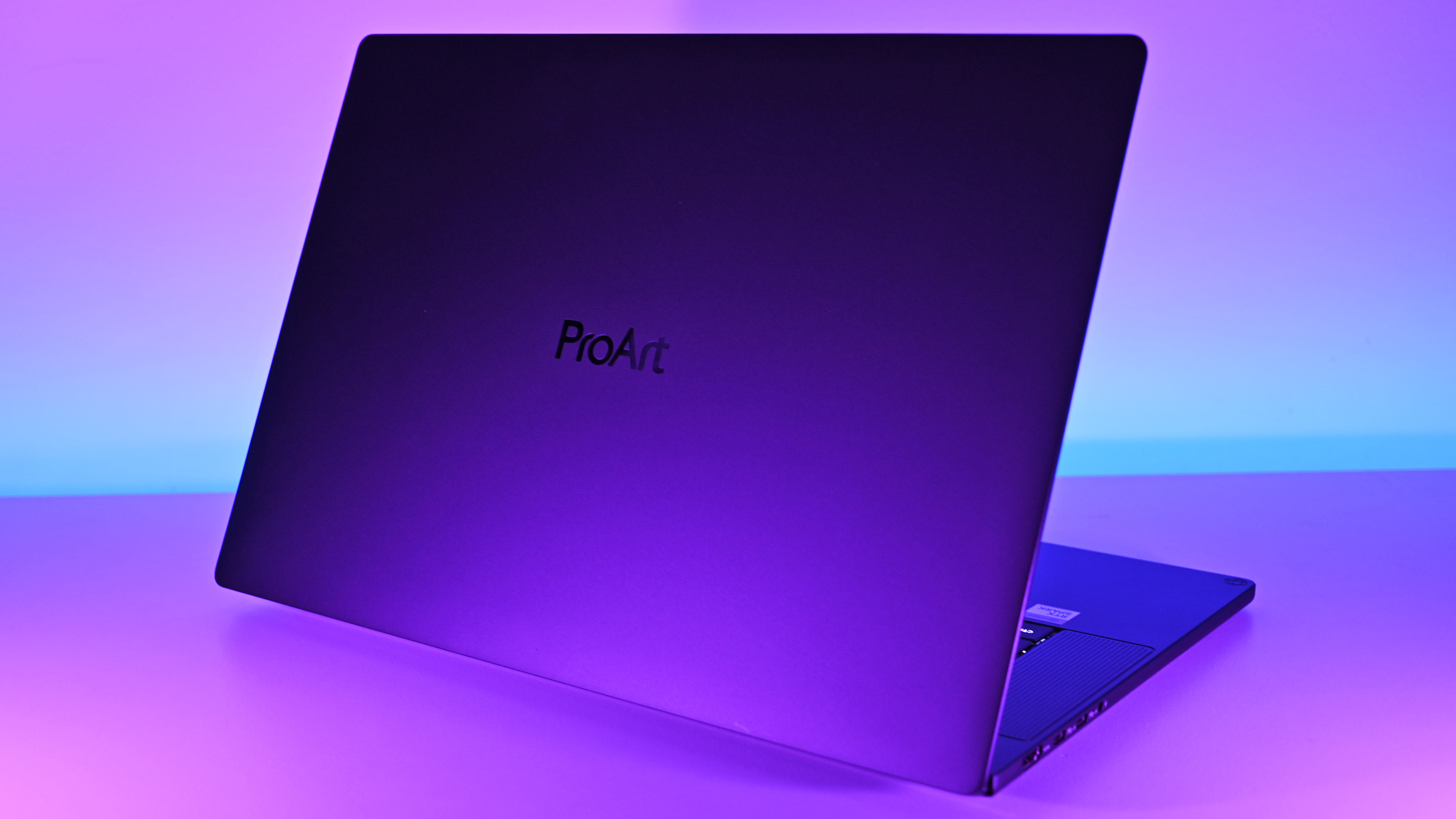 Rear angled view of the ASUS ProArt P16 laptop lid under vivid purple lighting, highlighting the centered glossy ProArt logo and slim profile.