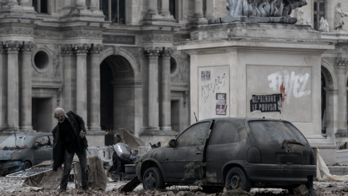 A Parisian walker dressed in a dark suit leers near a destroyed car and a graffitied monument in The Walking Dead: Daryl Dixon season 2