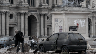 A Parisian walker dressed in a dark suit leers near a destroyed car and a graffitied monument in The Walking Dead: Daryl Dixon season 2