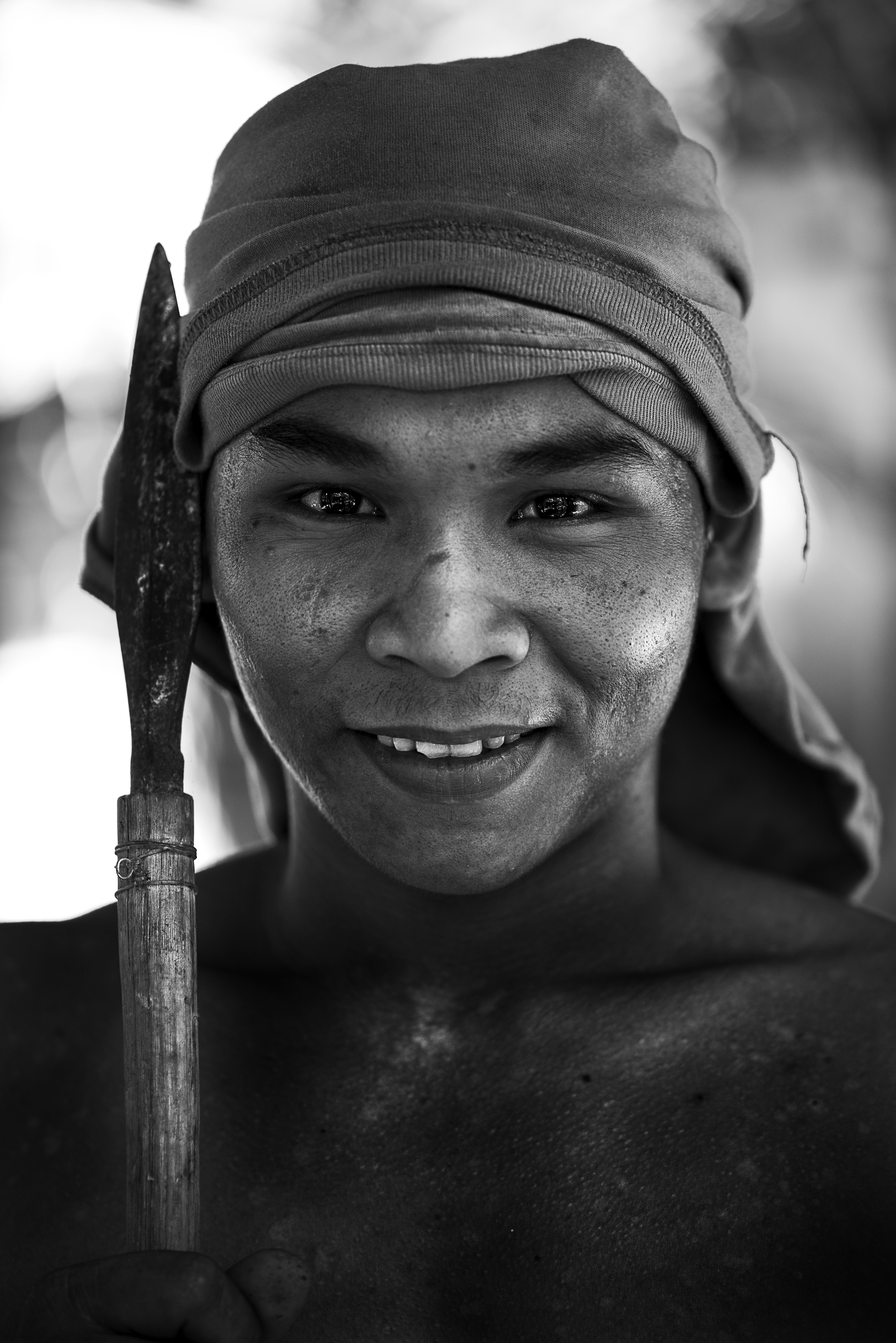 A black and white picture of a tribal man in Philippines wearing a shirt around his head and holding a spear next to his face. There is incredible sharpness and detail in his face and eyes