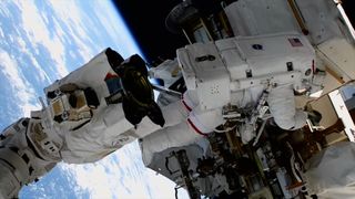 an astronaut in a white spacesuit works on a spacewalk near the truss of a space station and the end of a robotic arm