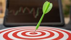 A dart sticks in the target of a dartboard while a tablet screen in the background shows a trading graph.