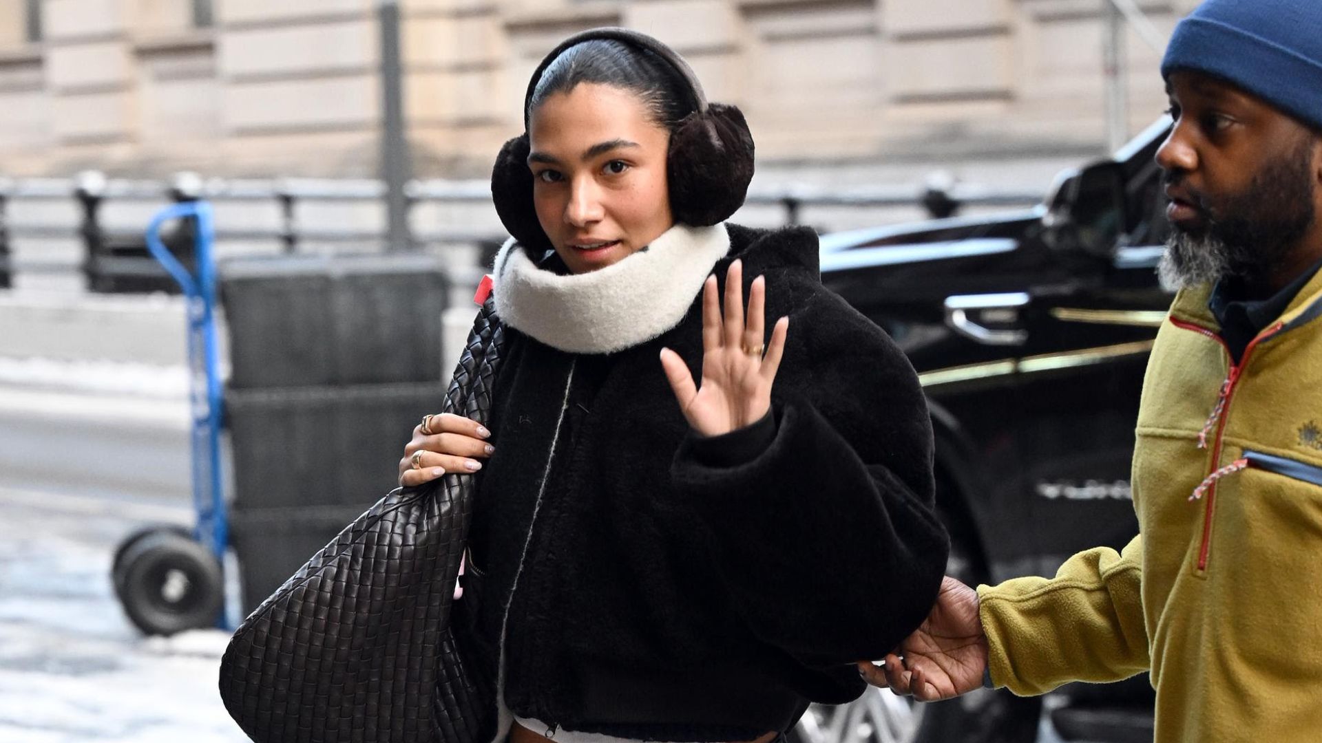 Olivia Dean walks outside wearing dark wash low-rise jeans with brown pointed-toe stiletto boots, a black bomber jacket, earmuffs and a Bottega Veneta Maxi Bag.