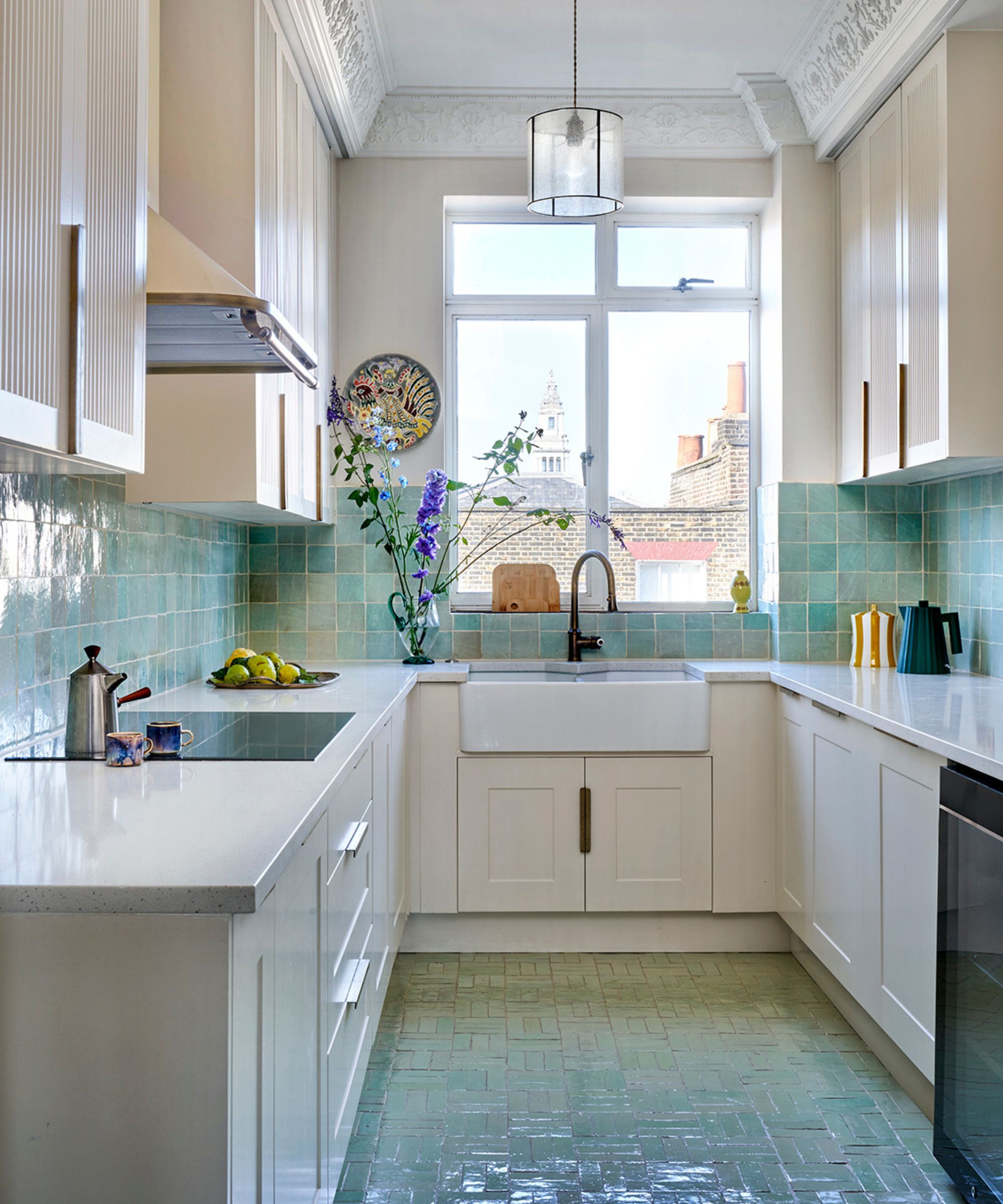 Bright galley kitchen with white cabinets, pale green tiled splashback and matching tiled floor. A large window above a farmhouse sink lets in daylight, highlighting simple worktops, plants, and soft pastel tones throughout the narrow space.