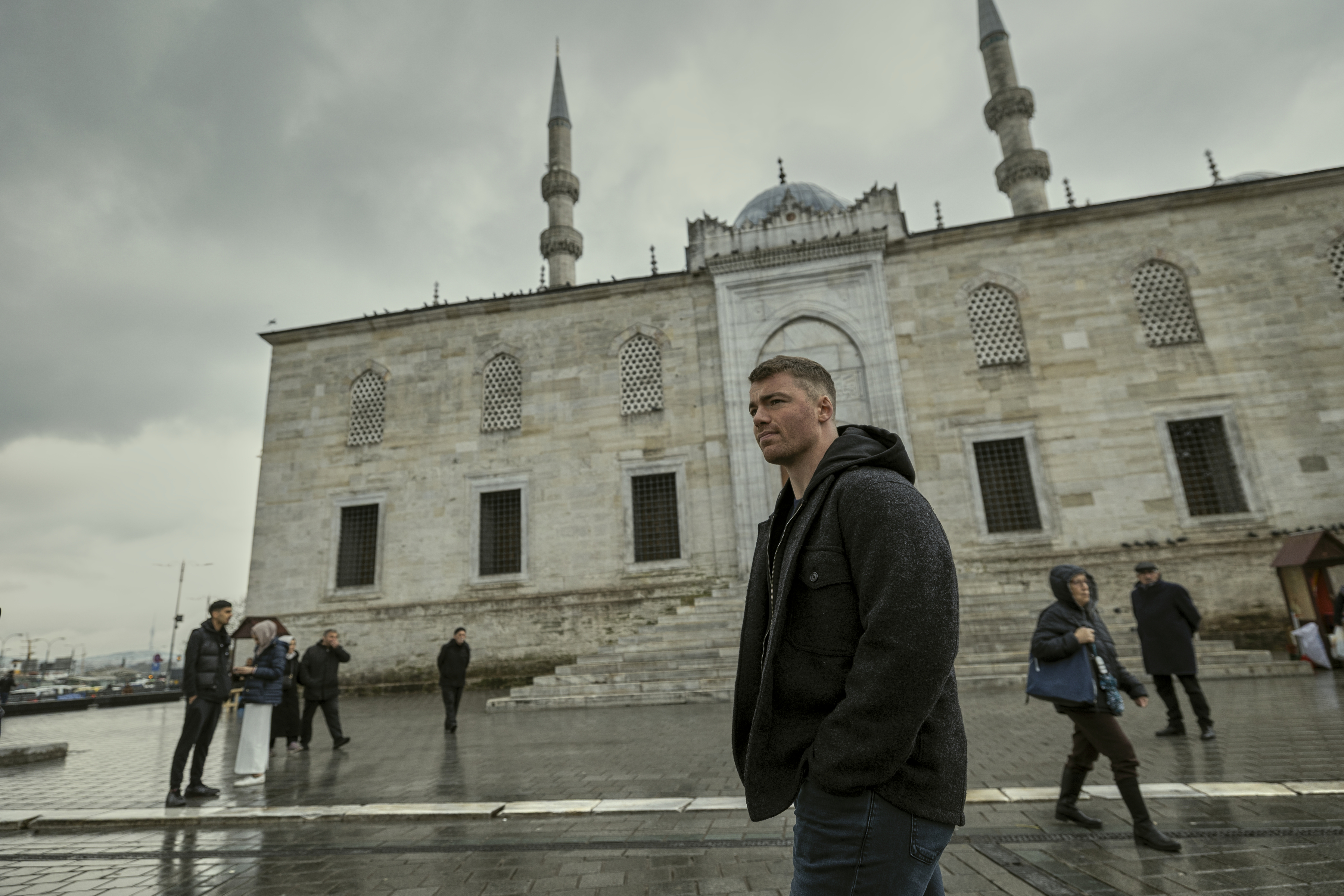 Gabriel Basso as Peter Sutherland walking through a rainy square by a river in the night agent season 3