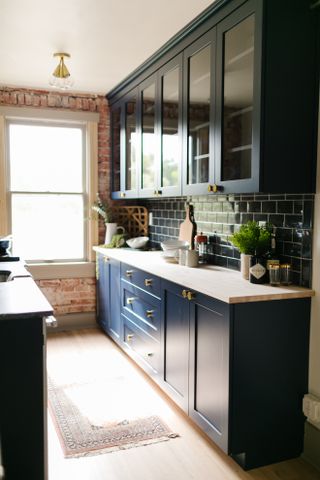 navy galley kitchen with wood countertops, brass knobs, window at end, navy metro tile splash back, navy wall cabinets with glass