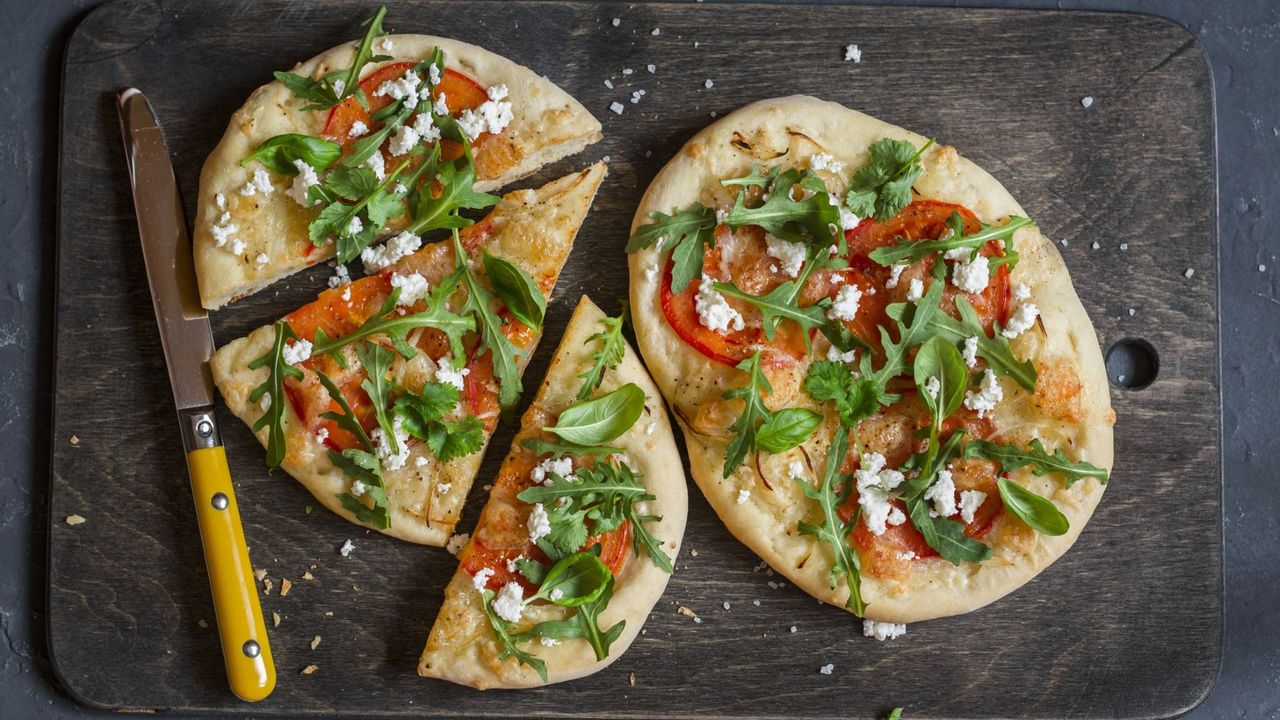 Two mini pizzas on a dark wood chopping board. One is cut, one is whole. They are topped with fresh tomatoes and arugula. 