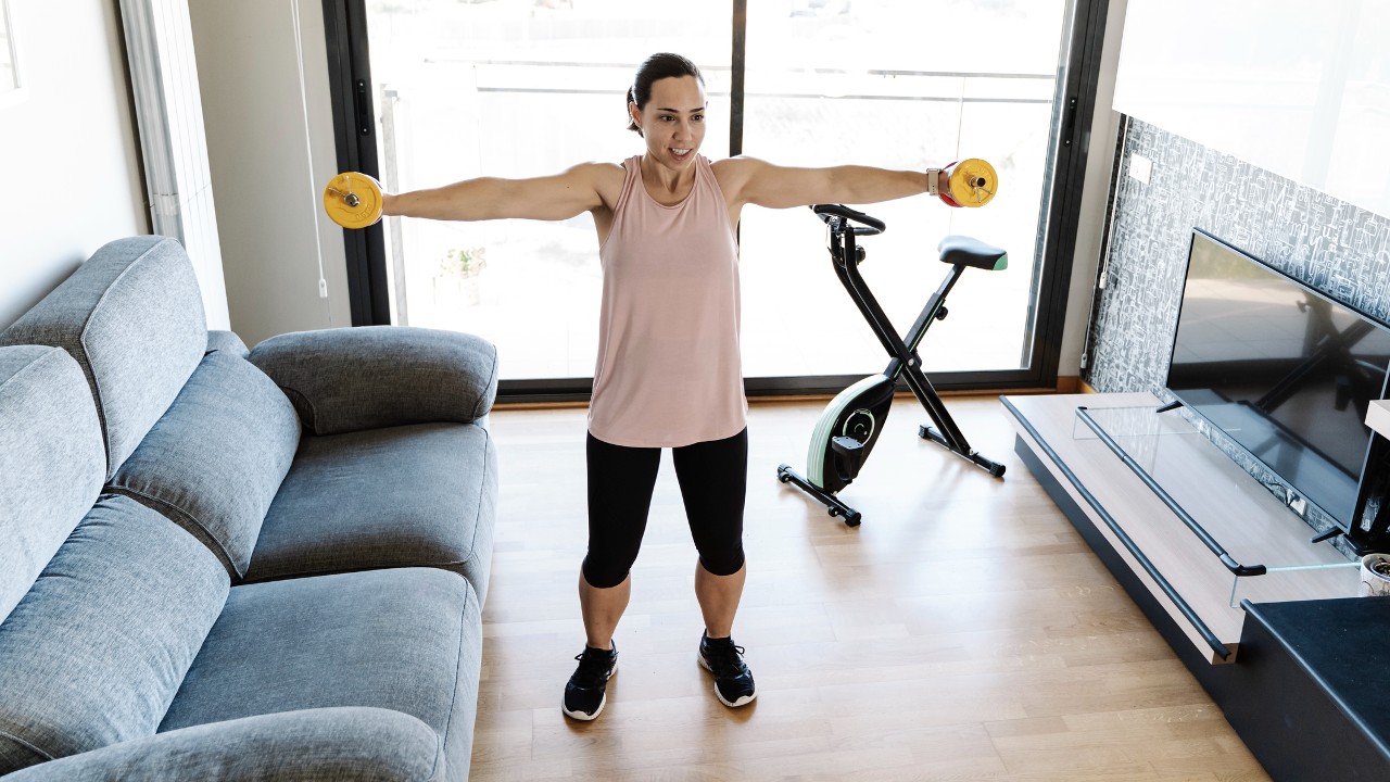Woman performs lateral raise shoulder exercise using dumbbells at home