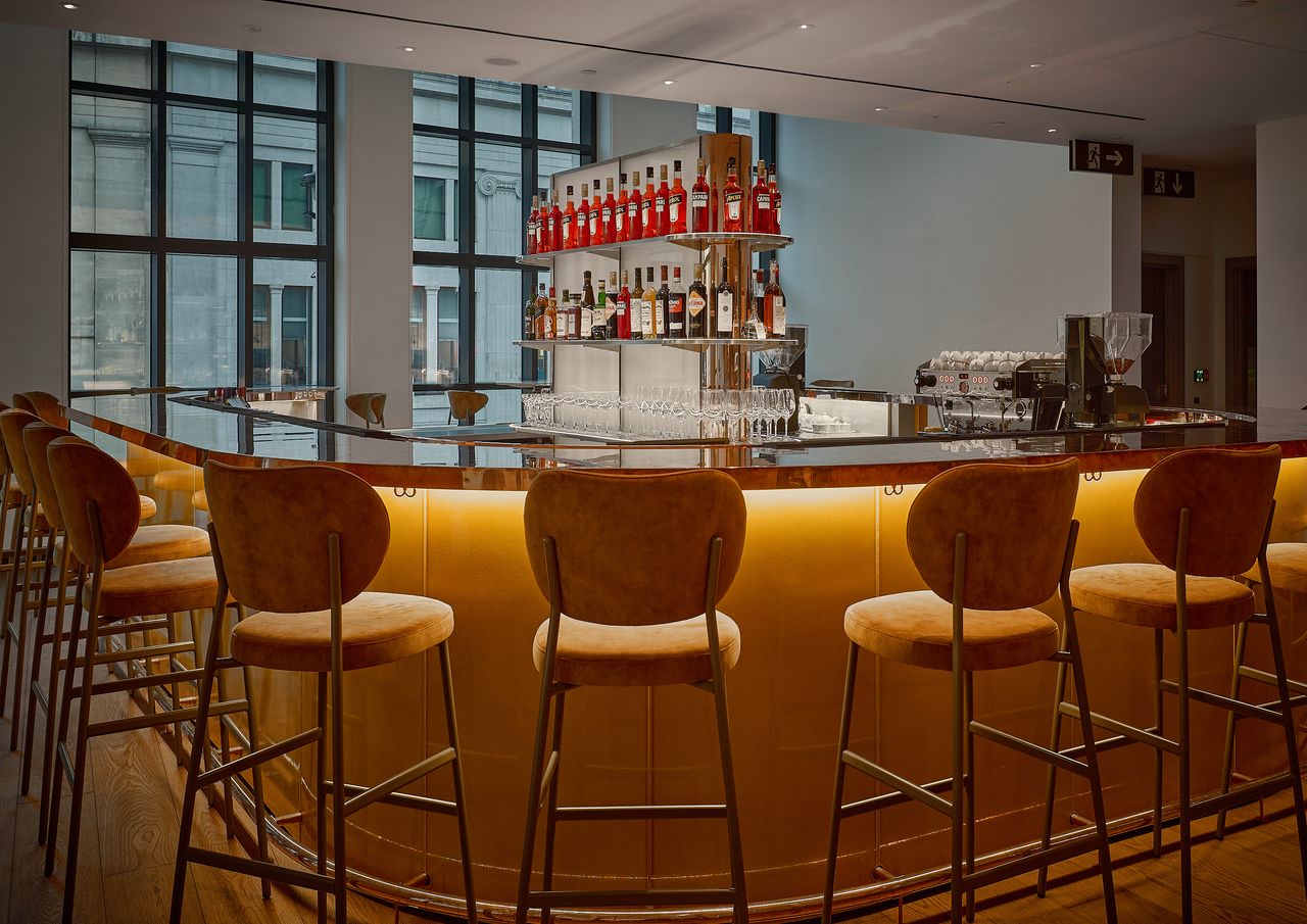 A dazzling cocktail bar features a sleek bronze countertop, encircled by velvety brown stools, and a central cabinette filled with all sorts of liqueurs.