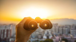 A person holds a pair of binoculars up to a sunset over a city in the distance. 