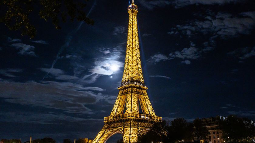 A view looking up at the Eiffel Tower lit in gold at night shining above the Paris skyline as the moon illuminates a bank of dappled clouds to the left of the steel structure.