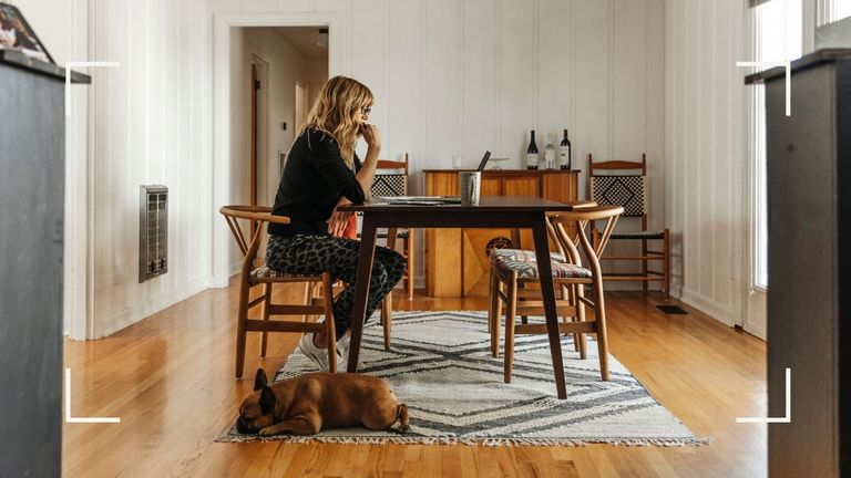Woman sitting at table looking at laptop at home with dog next to chair