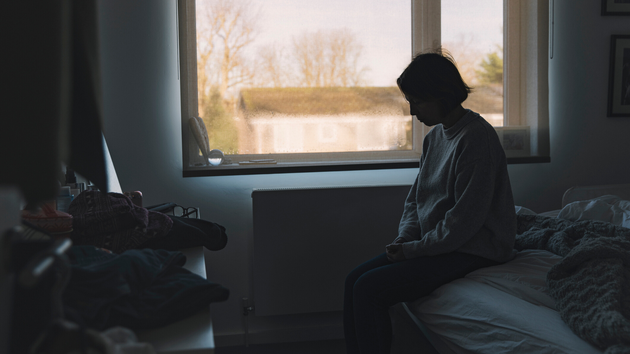 Woman sitting in a dark room with the window blinds open 
